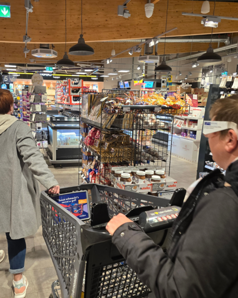 Eine Kundin mit einer Simualtionsbrille wird durch einen Supermarkt begleitet.