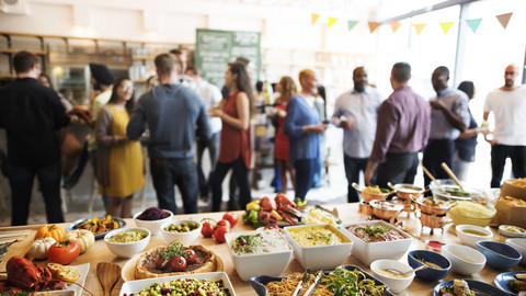 Im Vordergrund ist ein Buffet zu sehen. Im HIntergrund stehen Menschen