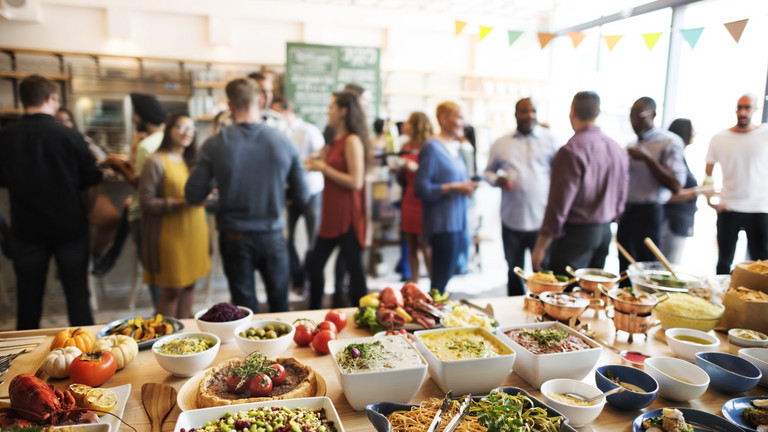 Das Bild zeigt eine Veranstaltung an, vorne das Buffet und im HIntergrund die Teilnehmer/innen. 