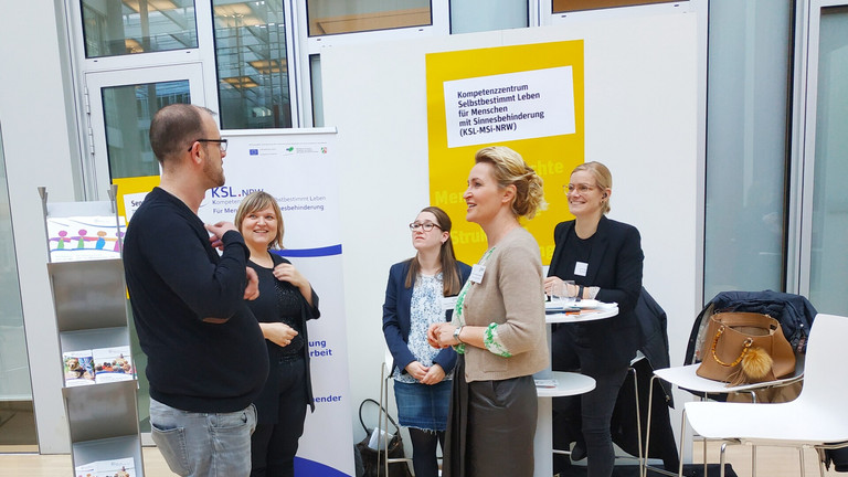 Es stehen fünf Personen am Messestand. Links steht ein Mann, hinten am Messestand sind Anna Cebulla, Melanie Wegerhoff und Kristin Reker. Vorne rechts steht Frau Janaszek und spricht mit dem Mann.