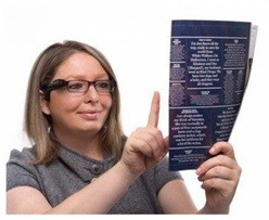 Eine Frau trägt die OrCam. Sie zeigt mit dem rechten Zeigefinder auf ein Dokument