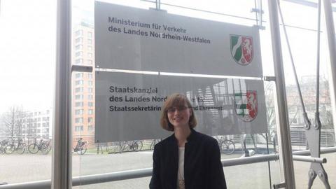 Frau Stiller steht vor dem Logo des Minsterium für Verkehr  des Landes Nordrhein Westfalen