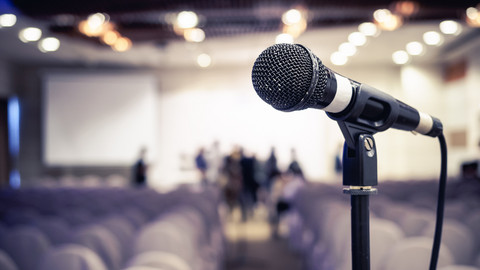 Ein Mikrophon steht auf einer Bühne. Im Hintergrund sind Stühle zu erkennen