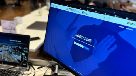 Computer-Bildschirm mit BodySigns-Homepage. Im Hindergrund ist ein verschwommendes Bild mit sitzenden Teilnehmenden an Tischen.