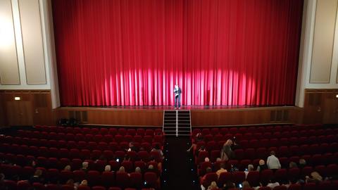 Ein Kinosaal mit rotem Vorhang. Ein Mann steht auf der Bühne und spricht in ein Mikrofon