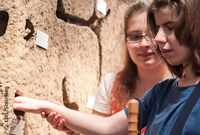 Ein blindes Mädchen ertastet ein lebensgroßes Modell. Ihre Hand wird von einem anderen Mädchen geführt.