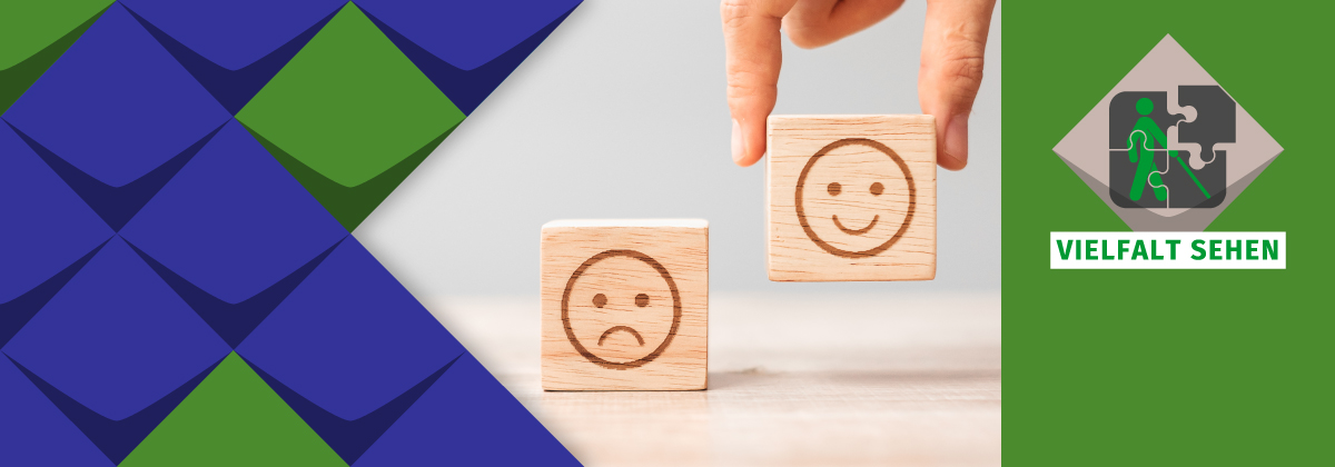 Ein Holzbaustein auf einer Tischplatte, darauf ein trauriges Gesicht. Ein weiterer Baustein wird von oben abgesetzt. Auf ihm ein fröhliches Gesicht.