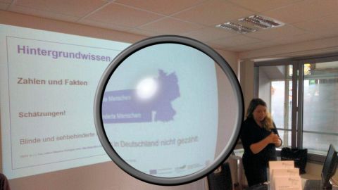 Ramona Armbrust steht vor einer Wand auf der eine Bildschirmpräsentation ist. Auf der Folie steht "Hintergrundwissen, Zahlen und Fakten". In der Mitte des Bildes ist eine große Lupe eingefügt, die einen Teil des Fotos vergrößert.