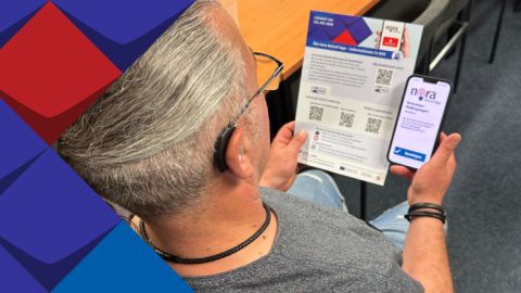 Ein Mann mit dem Hörgerät hat ein Smatphone mit dem Logo nora Notruf-App und liest ein Infoblatt zum Thema nora Notruf-App